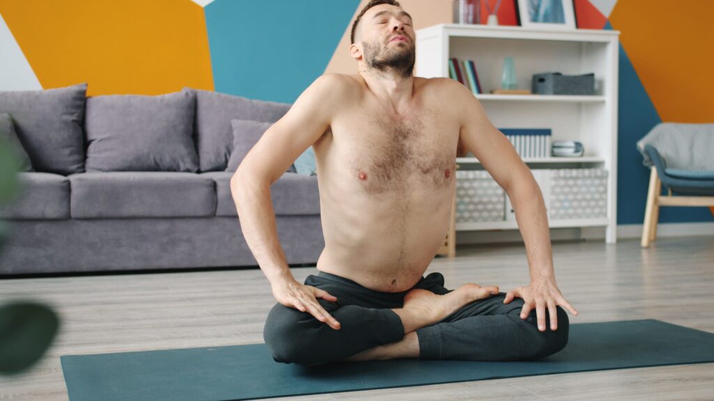 Breathing Techniques in Yoga: The Foundation of Physical and Mental Well-Being Shirtless man performing yoga meditation pose on mat in modern living room.