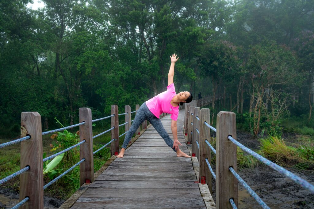 Full body of fit female in activewear on wooden footbridge doing Triangle pose while exercising asana in nature with trees